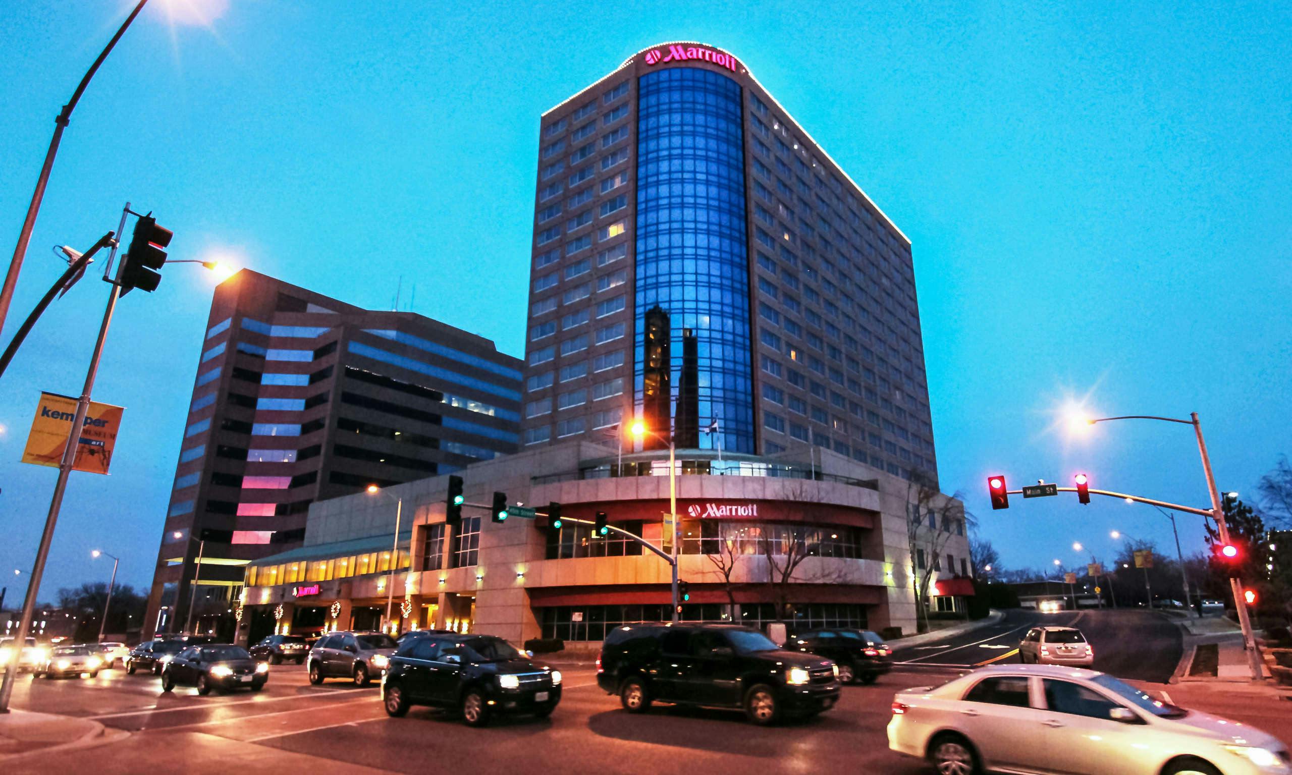 Image source: https://www.hoteltonight.com/hotel/marriott-country-club-plaza-in-kansas-city-mo Marriott Country Club Plaza hotel - Marriott hotel tower rises above a busy city intersection at dusk, with illuminated signage, traffic lights and vehicles moving below.