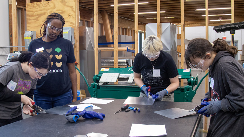 Participants wearing safety glasses and gloves cut sheet metal at a worktable, with industrial equipment visible in the lab behind them.