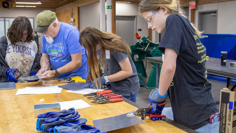 Participants wearing safety glasses and gloves use hand tools to cut sheet metal while working around a shop table in a trades lab.